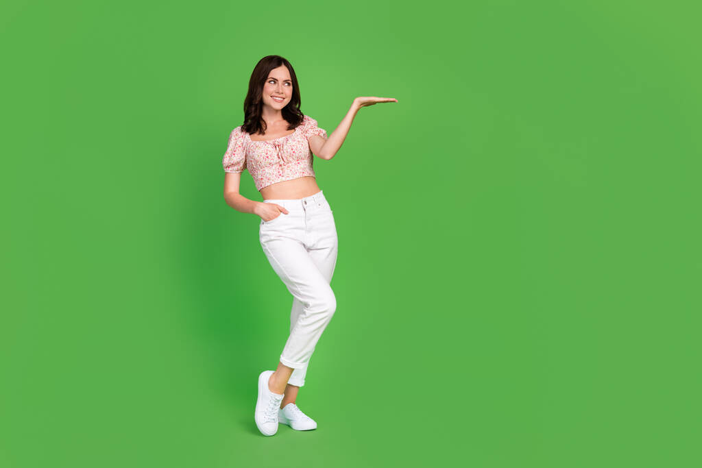 Foto completa di splendida bella ragazza indossare camicetta pantaloni bianchi mano in tasca presentando oggetto isolato su sfondo di colore verde. - Foto, immagini