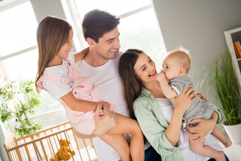 Photo de mignon couple positif souriant serrant bras tenant deux enfants à l'intérieur de la maison chambre à la maison. - Photo, image