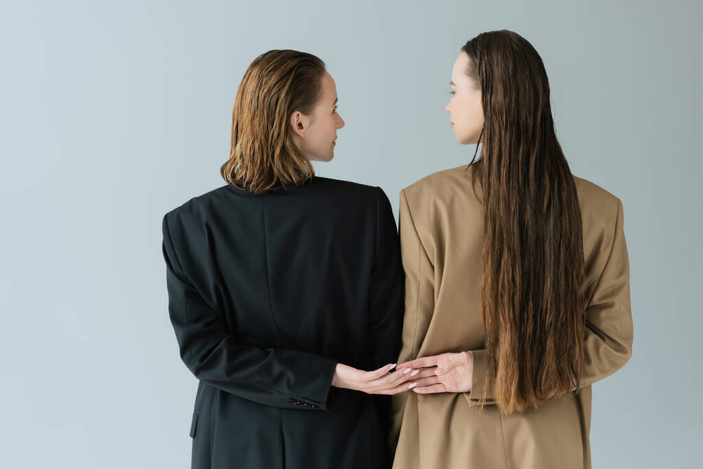 back view of lesbian women in black and beige jackets looking at each other and holding hands isolated on grey - Photo, Image