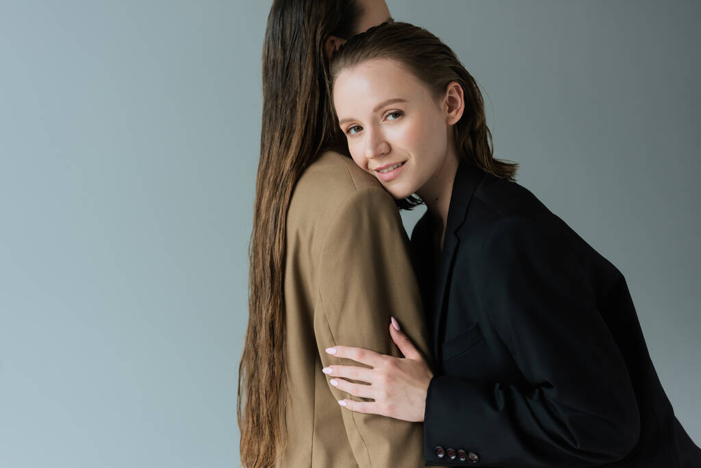 mujer lesbiana complacida sonriendo a la cámara y abrazando a su novia con el pelo largo aislado en gris - Foto, Imagen