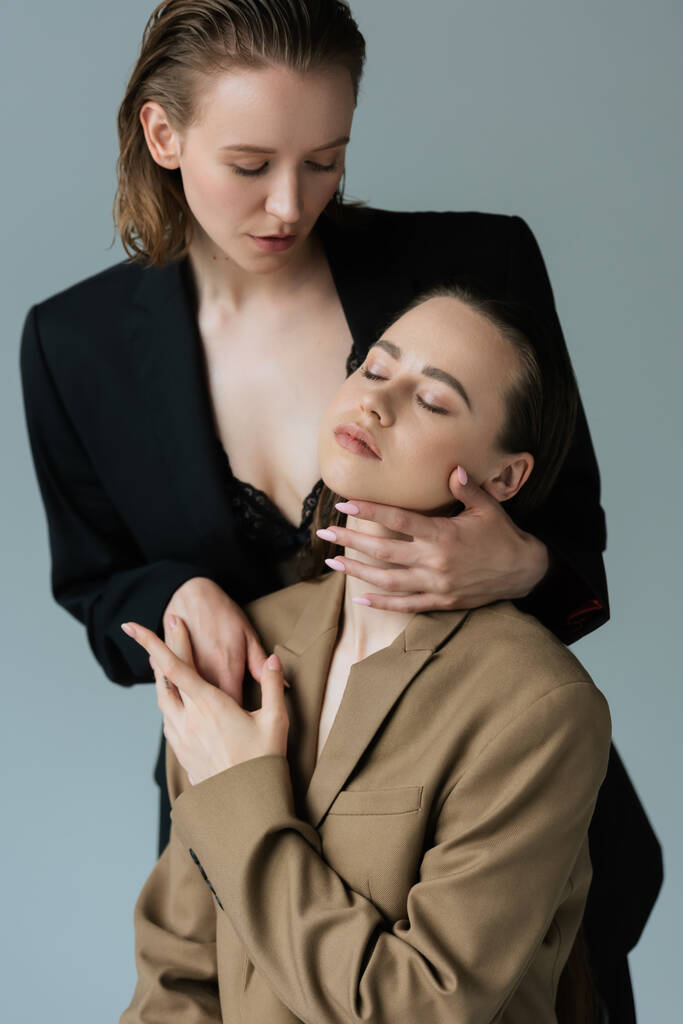 сексуальная лесбиянка в черном лифчике и блейзере, соблазняющая молодую девушку, изолированную от серых - Фото, изображение