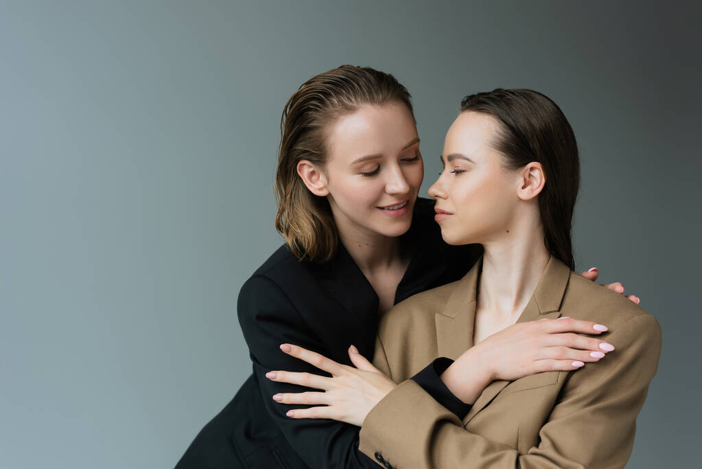mosolygó leszbikusok bézs és fekete blézer ölelés elszigetelt szürke - Fotó, kép