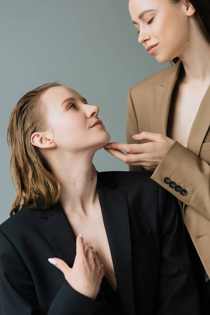 fiatal nő bézs blézer megérinti áll fiatal leszbikus barátnő elszigetelt szürke - Fotó, kép