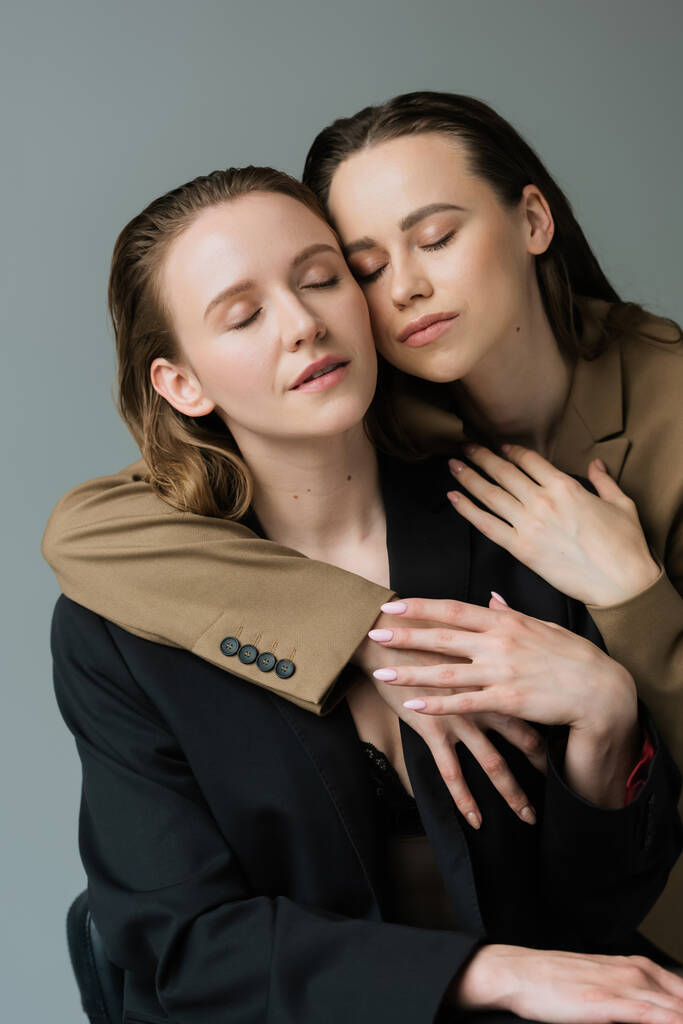 couple sensuel de lesbiennes en blazers beige et noir embrassant les yeux fermés isolé sur gris - Photo, image