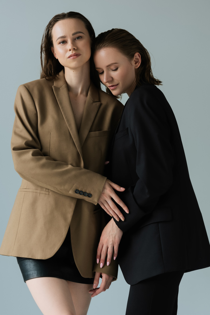brunette woman in beige blazer and black skirt looking at camera near lesbian partner isolated on grey - Photo, Image