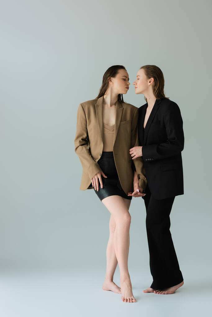γυναίκα με μαύρο κοστούμι και η λεσβία σύντροφός της με σακάκι και φούστα κρατώντας τα χέρια και κοιτάζοντας ο ένας τον άλλον σε γκρι φόντο - Φωτογραφία, εικόνα