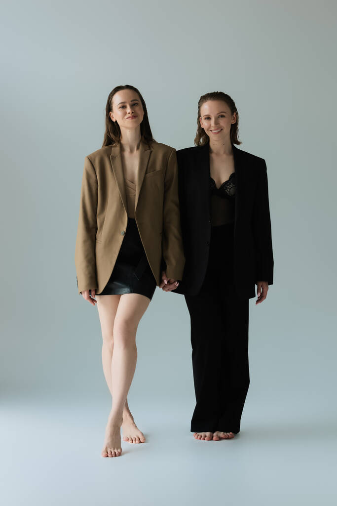 barefoot woman in blazer and skirt and her lesbian girlfriend in suit holding hands and smiling at camera on grey  - Photo, Image