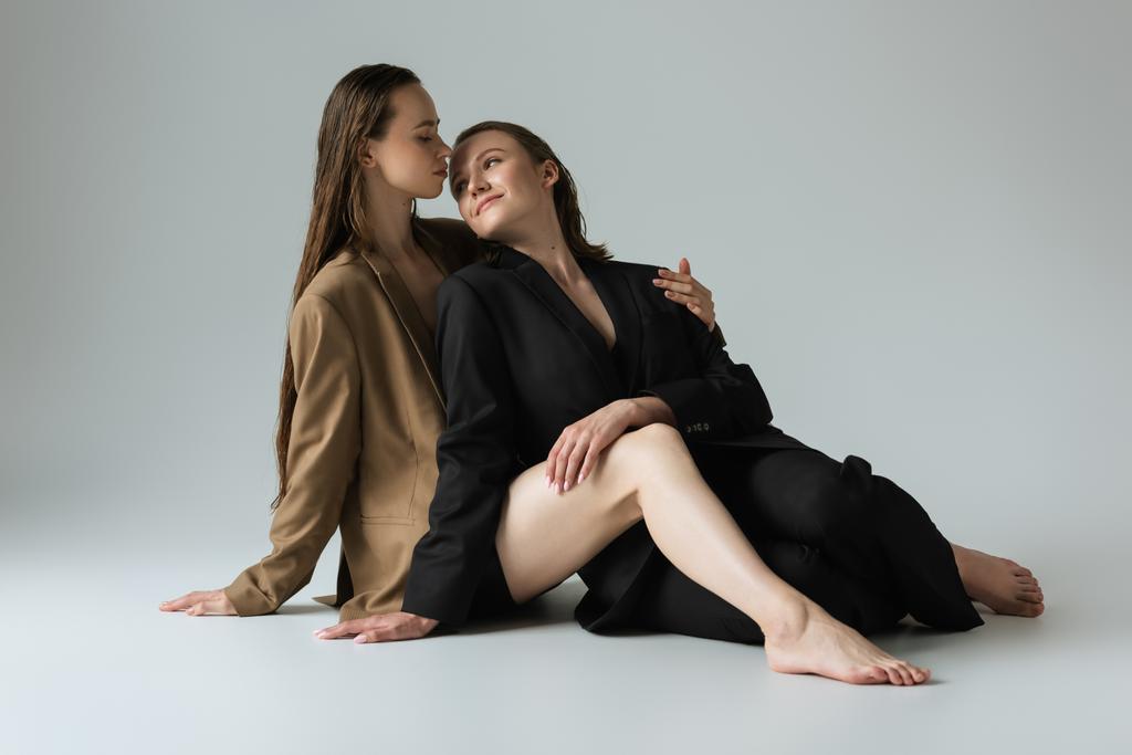 Barfüßige lesbische Frauen in Blazern lächeln einander an, während sie auf grauem Hintergrund sitzen - Foto, Bild