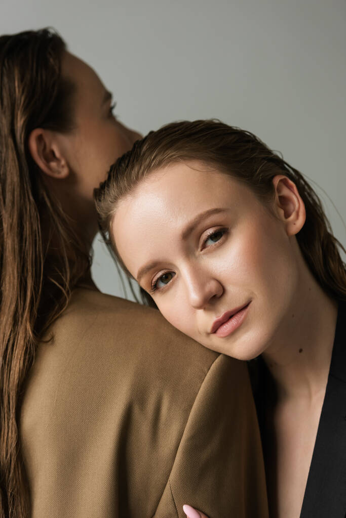 young lesbian woman looking at camera while leaning on girlfriend isolated on grey - Photo, Image
