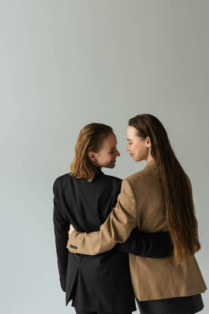 vista trasera de pareja del mismo sexo en chaquetas abrazando y sonriendo aislado en gris - Foto, Imagen
