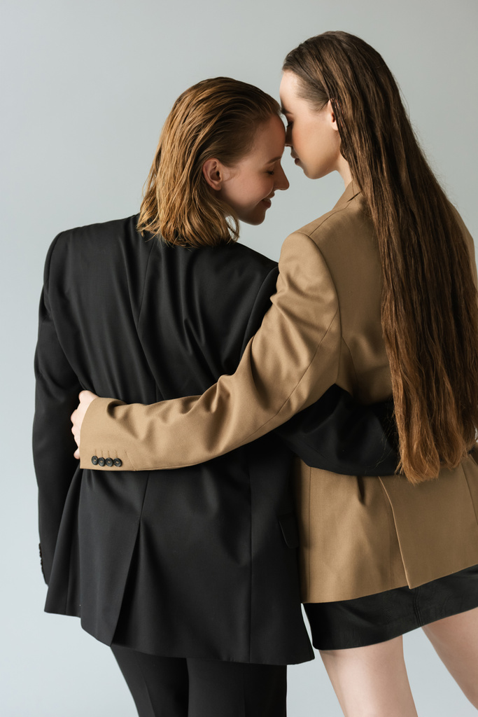 πίσω όψη της γυναίκας με μακριά μαλλιά αγκαλιάζει νεαρή λεσβία φίλη απομονωμένη σε γκρι - Φωτογραφία, εικόνα