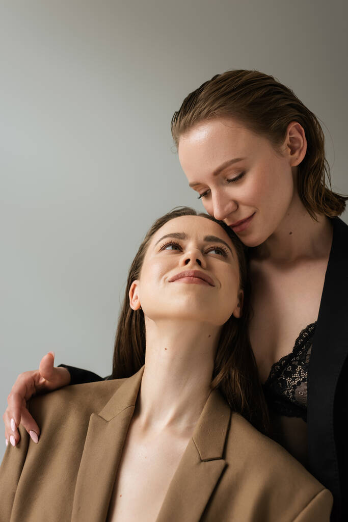 красивая лесбиянка, улыбающаяся соблазнительной подружке-лесбиянке в черном лифчике и пиджаке, изолированном от серых - Фото, изображение