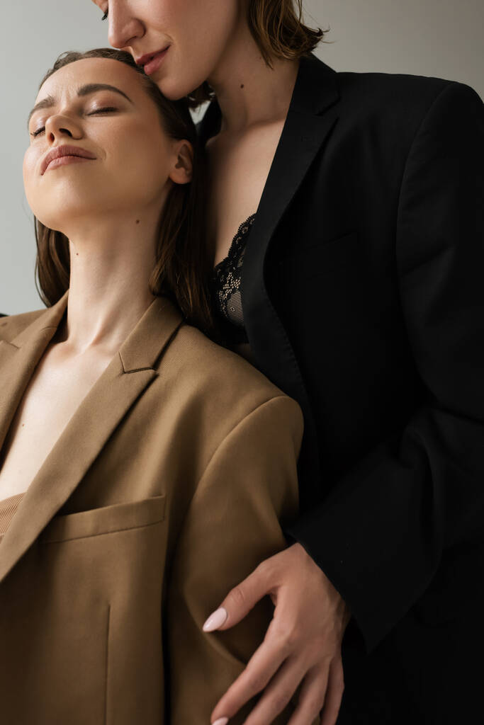 γυναίκα σε μπεζ σακάκι στέκεται με κλειστά μάτια κοντά λεσβία φίλη αγκαλιάζει της απομονωμένη σε γκρι - Φωτογραφία, εικόνα