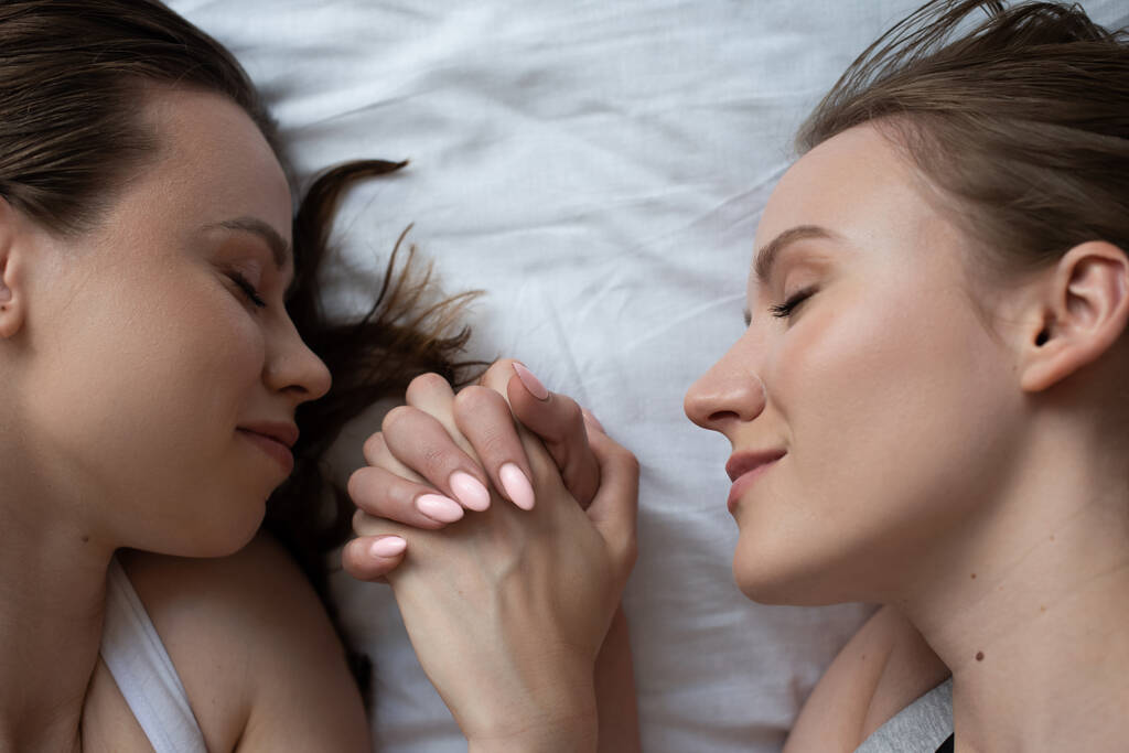 vista superior de amigas lesbianas felices tumbadas sobre ropa de cama blanca con los ojos cerrados y cogidas de la mano - Foto, Imagen