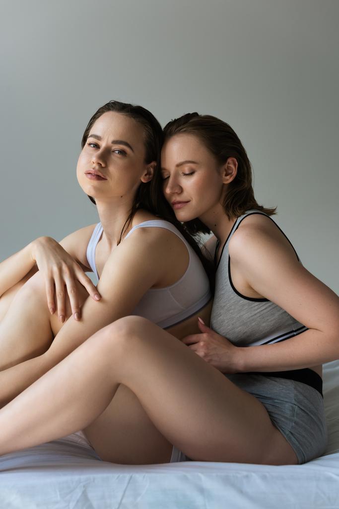 sinnliche lesbische Frau blickt in die Kamera, während sie in der Nähe einer jungen Freundin auf grau isoliert sitzt - Foto, Bild