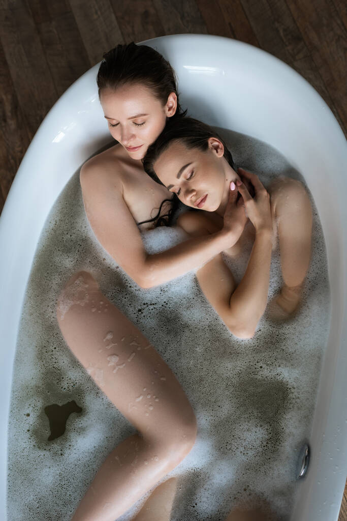 top view of nude lesbian couple with closed eyes taking bubble bath at home - Photo, Image