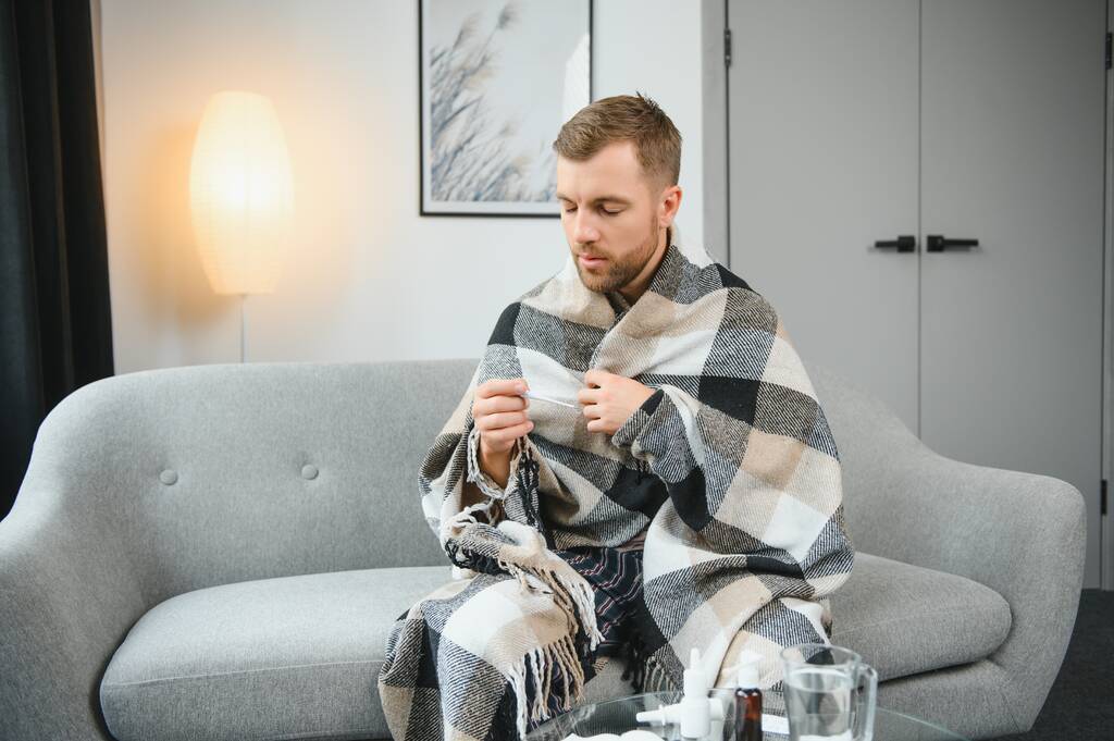 Chory brodaty mężczyzna, który ma przeziębienie lub grypę sezonową, siedzący na kanapie w domu. Gość z gorączką, noszący ciepłą kratę, drżący ze zmartwienia. - Zdjęcie, obraz
