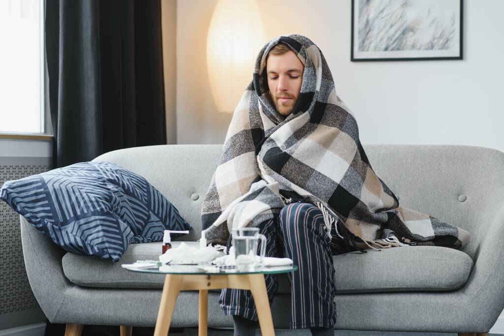 Ένας άρρωστος άνθρωπος κάθεται στο σπίτι σε ένα γκρι καναπέ με μια κουβέρτα. Ασθένεια, προστασία, coronavirus, ασθένεια, γρίπη. - Φωτογραφία, εικόνα