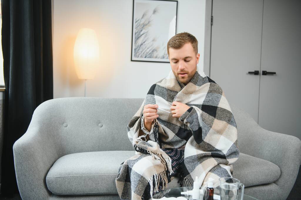 Kranker Mann liegt auf Sofa und überprüft seine Temperatur unter einer Decke zu Hause im Wohnzimmer. - Foto, Bild