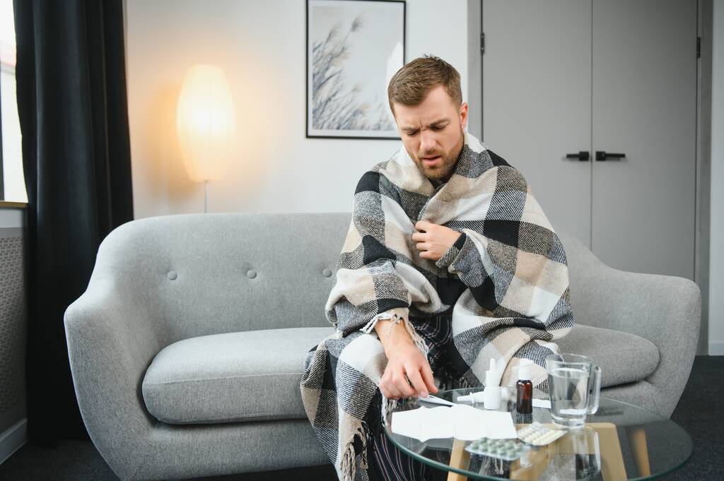 病人がリビング ルームで自宅で毛布の下彼の温度をチェックするソファに横たわっています。. - 写真・画像