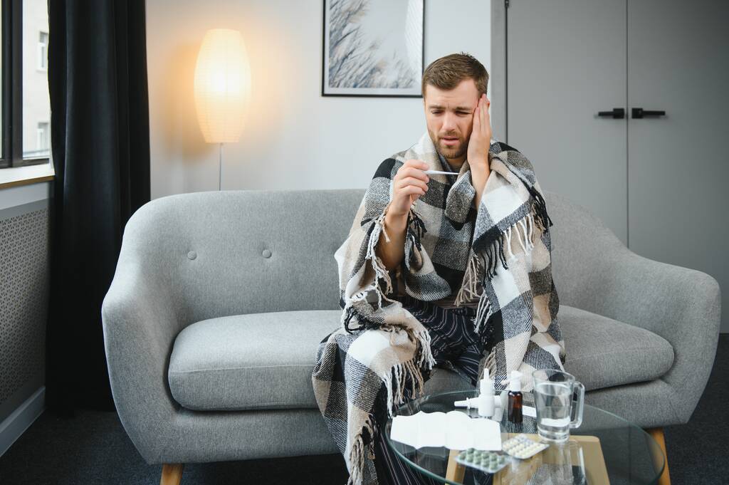 Malade homme barbu qui a mauvais rhume ou grippe saisonnière assis sur le canapé à la maison. Le gars avec de la fièvre portant des frissons à carreaux chauds avec expression inquiète du visage - Photo, image