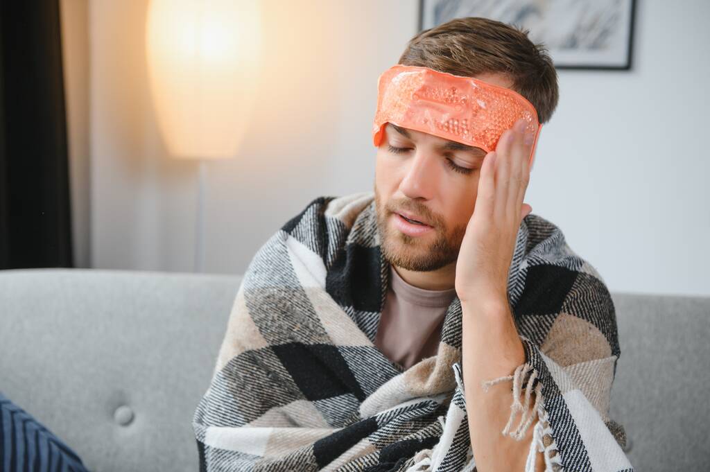 Άρρωστος γενειοφόρος που έχει κρυολογήματα ή εποχική γρίπη και κάθεται στον καναπέ στο σπίτι. Ο τύπος με τον πυρετό φοράει ζεστό καρό ρίγος με ανήσυχη έκφραση προσώπου - Φωτογραφία, εικόνα