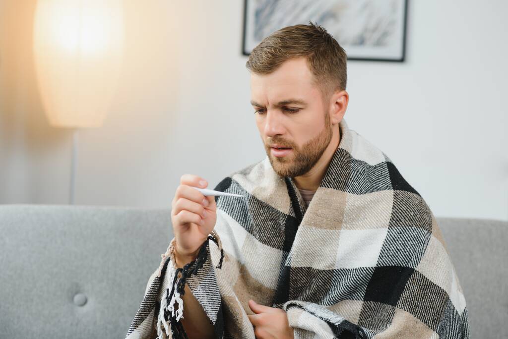 Kranker erwachsener Mann mit schwerer Erkältung oder saisonalem Grippevirus kämpft zu Hause gegen Krankheit. Unzufriedener Mann mittleren Alters sitzt auf Sofa, in warmen Schal und Karo gehüllt, hält Papiertaschentücher in der Hand und niest - Foto, Bild