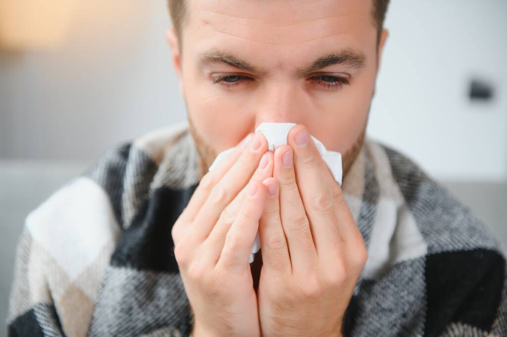 Hombre joven que sufre de un resfriado común y gripe o alergia se sienta en casa envuelto en manta y se limpia la nariz con tejidos mientras tiene un fuerte dolor de cabeza, concepto de atención médica - Foto, Imagen