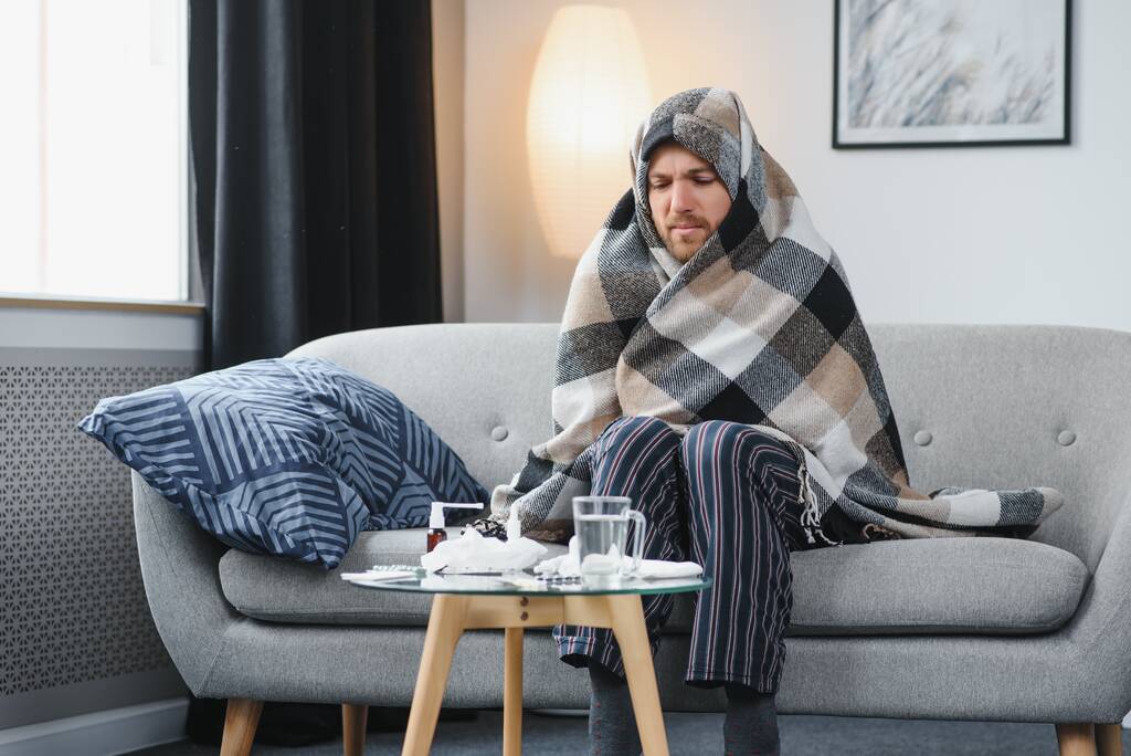 Een zieke man zit thuis op een grijze bank met een deken. Ziekte, bescherming, coronavirus, ziekte, griep. - Foto, afbeelding