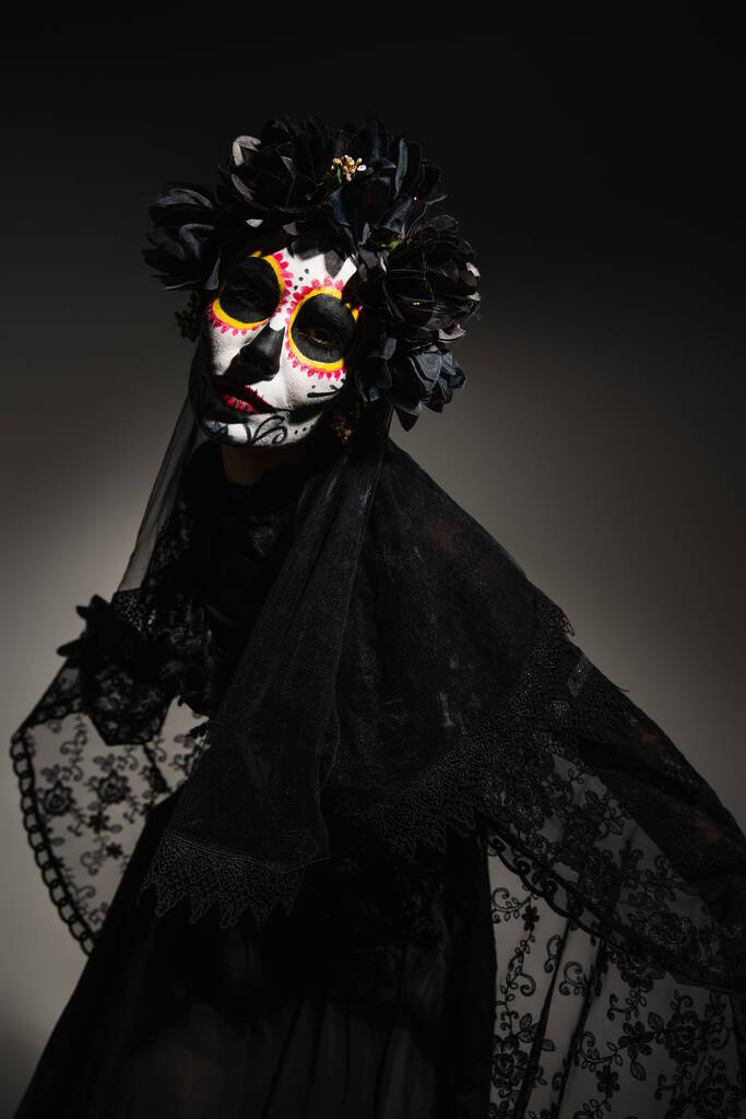 Frau mit Catrina-Make-up und dunklem Kranz auf dem Kopf blickt in die Kamera auf schwarzem Hintergrund  - Foto, Bild