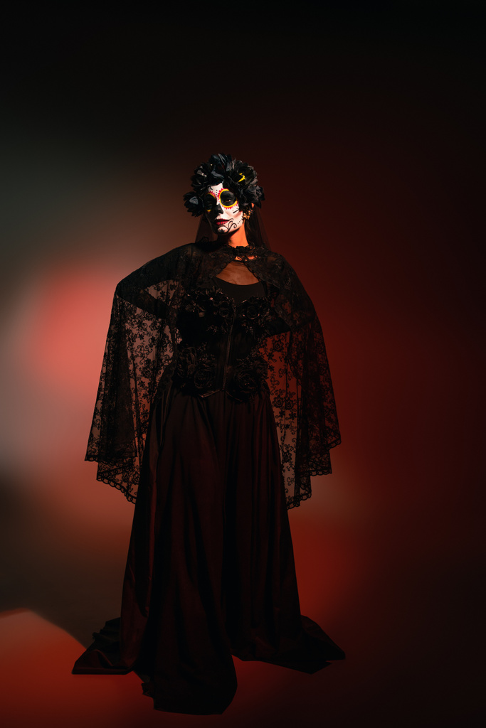 Γυναίκα σε μαύρο κοστούμι και ανατριχιαστικό αποκριάτικο μακιγιάζ στέκεται σε μπορντό φόντο  - Φωτογραφία, εικόνα