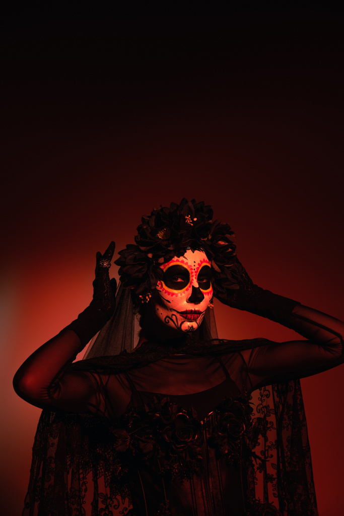 Woman in santa muerte traditional costume adjusting wreath on burgundy background with red lighting  - Photo, Image