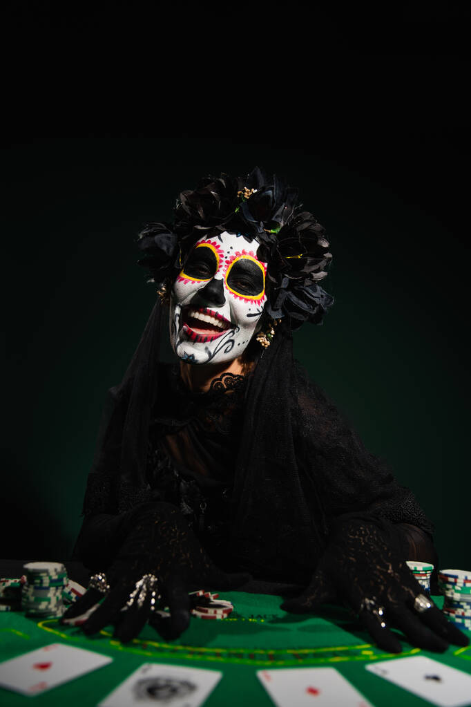 KYIV, UCRAINA - 12 SETTEMBRE 2022: Donna sorridente in costume da Santa Muerte vicino a giocare patatine e carte sul verde scuro - Foto, immagini