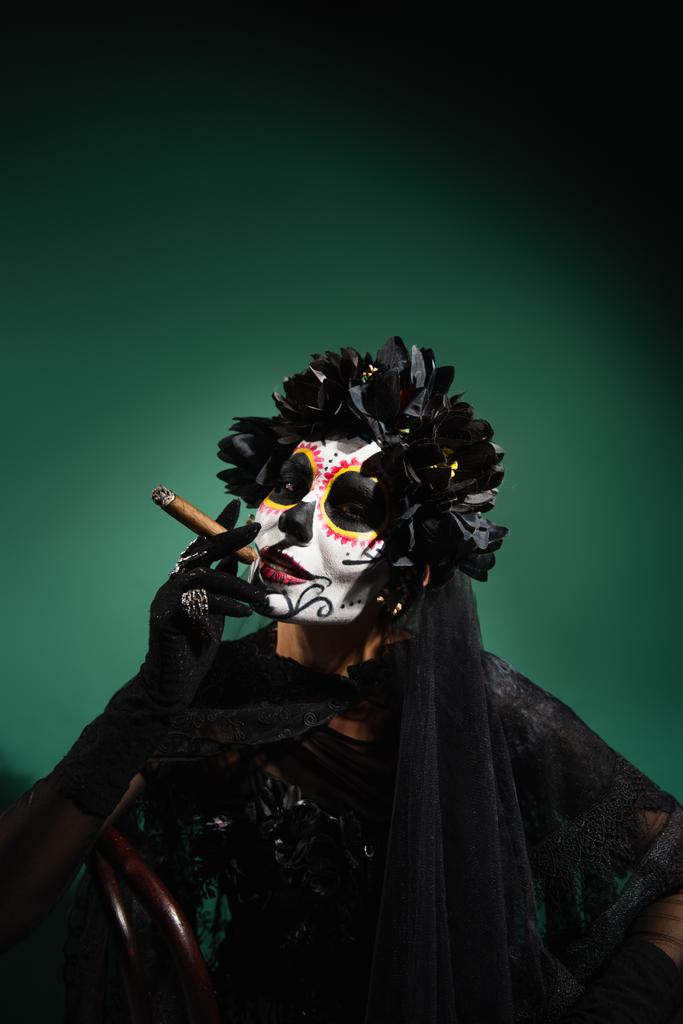 Woman in day of death makeup and black costume smoking cigar on green background  - Photo, Image