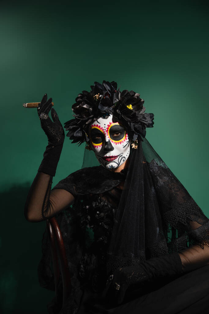 Γυναίκα στο μεξικάνικο κοστούμι Σάντα Μουέρτε και μακιγιάζ κρατώντας πούρο και κοιτάζοντας κάμερα στο πράσινο φόντο  - Φωτογραφία, εικόνα