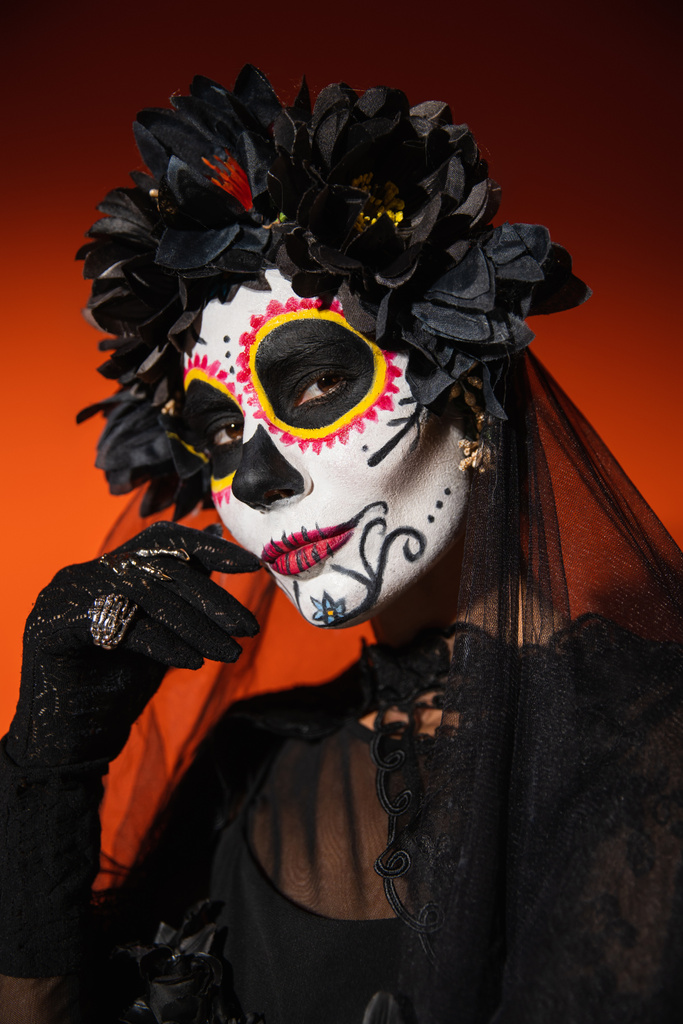 nő fekete koszorú és kísérteties halloween smink fogja kezét az arc mellett, és nézi a kamera narancssárga háttér - Fotó, kép