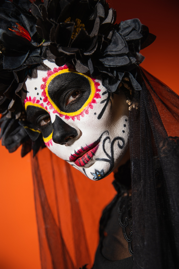 portrait of woman in traditional mexican day of dead makeup and black wreath looking at camera on orange background - Photo, Image