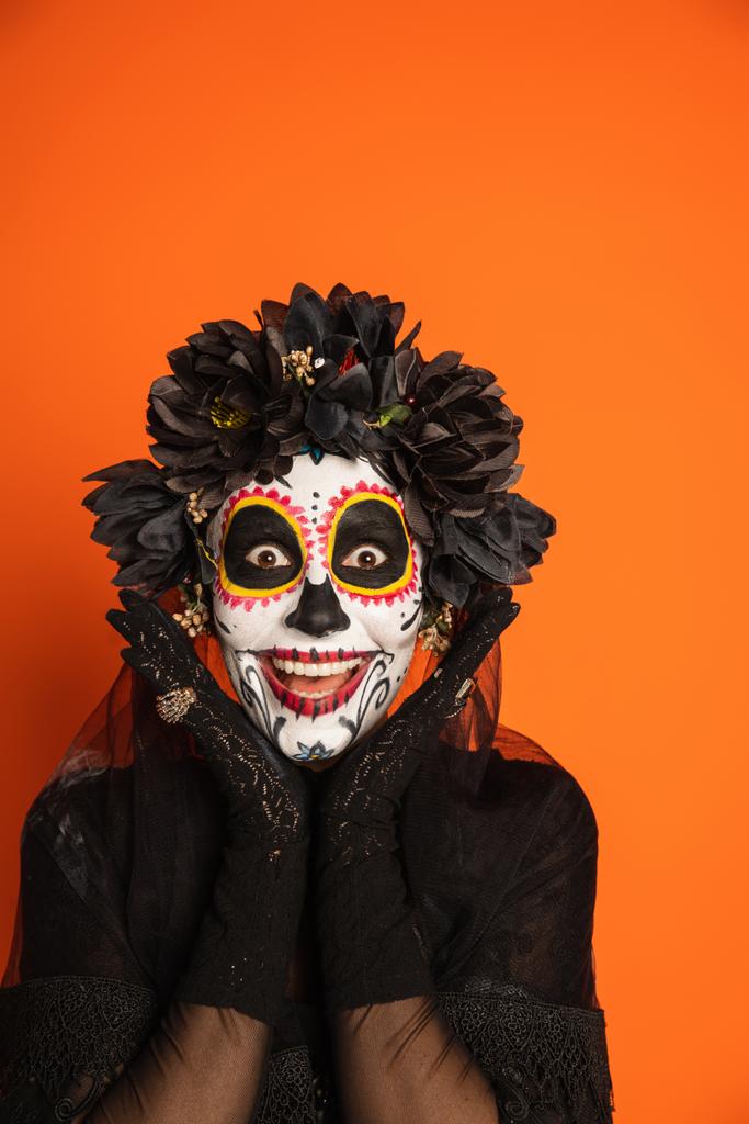 animado mulher em assustador halloween maquiagem e grinalda preta de mãos dadas perto do rosto e olhando para a câmera isolada em laranja - Foto, Imagem