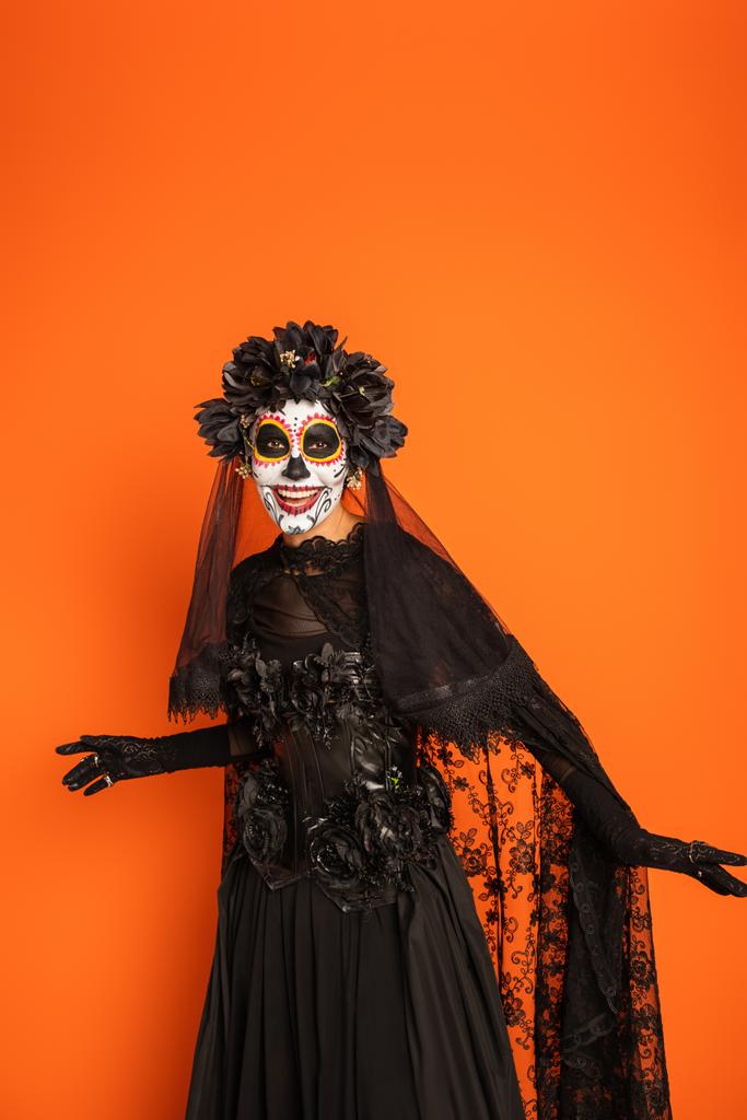 mulher sorridente em vestido de bruxa preta e assustador halloween maquiagem isolada em laranja - Foto, Imagem