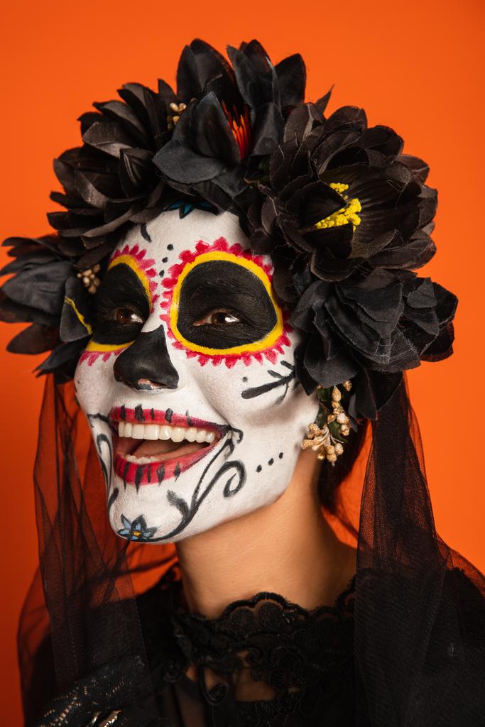 portrait of joyful woman in scary halloween makeup and black wreath with veil isolated on orange - Photo, Image