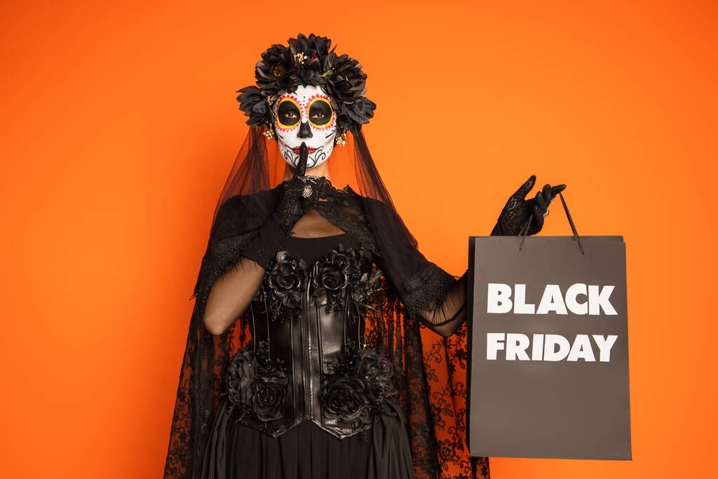 vrouw in suiker schedel halloween make-up en kostuum tonen stilte en het houden van zwarte vrijdag boodschappentas geïsoleerd op oranje - Foto, afbeelding