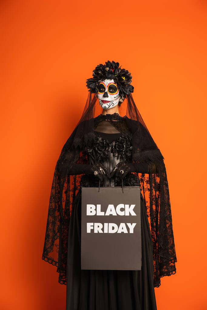 mujer en maquillaje catrina y traje negro con velo sosteniendo bolso de compras viernes negro aislado en naranja - Foto, Imagen
