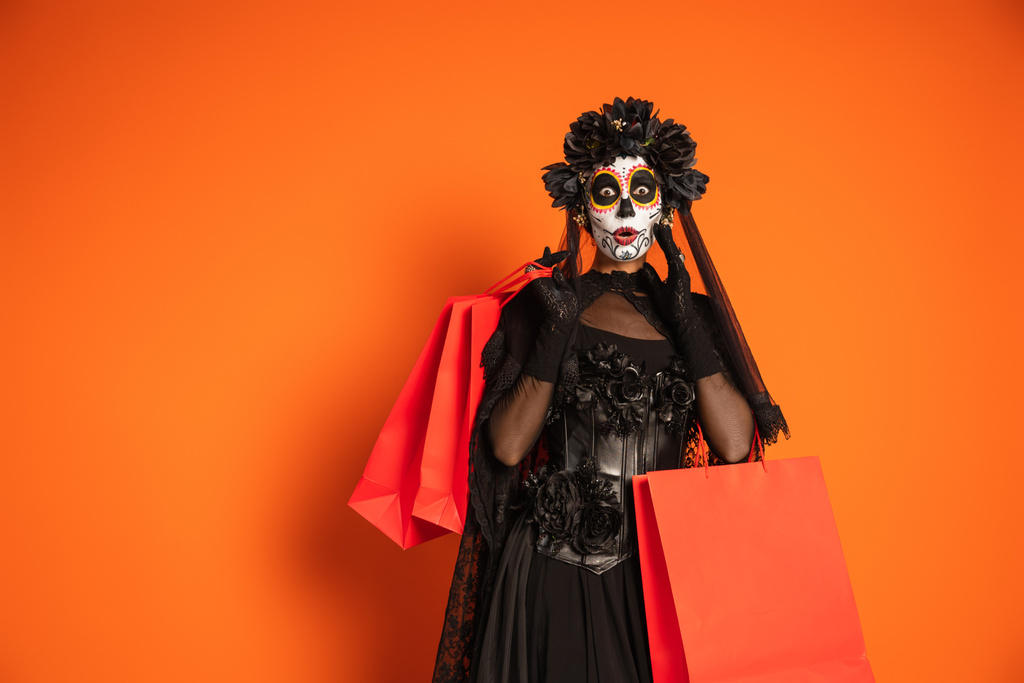 mulher surpreso em assustador halloween maquiagem e preto fantasia segurando sacos de compras no fundo laranja - Foto, Imagem