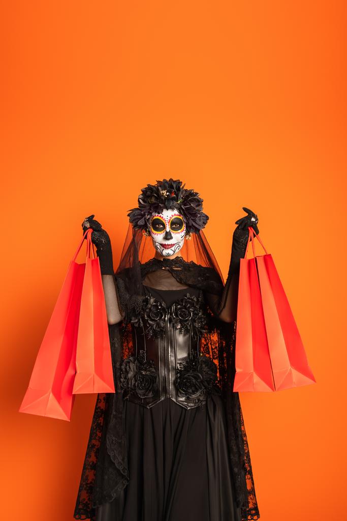 γυναίκα σε catrina μακιγιάζ και ανατριχιαστικό μαύρο κοστούμι με στεφάνι δείχνει τσάντες ψώνια που απομονώνονται σε πορτοκαλί - Φωτογραφία, εικόνα