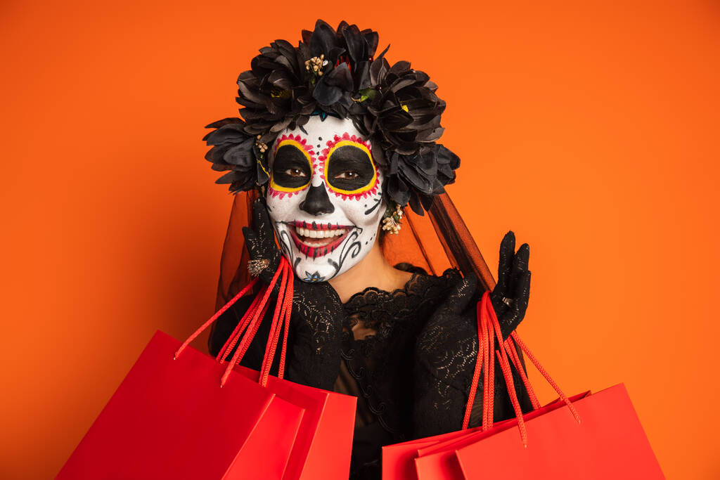 mujer alegre en corona negra y el cráneo de azúcar de Halloween maquillaje mirando a la cámara cerca de bolsas de compras aisladas en naranja - Foto, imagen