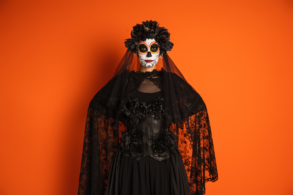 mujer en maquillaje tradicional de santa muerte y traje con velo de encaje negro de pie con las manos detrás de la espalda aisladas en naranja - Foto, imagen