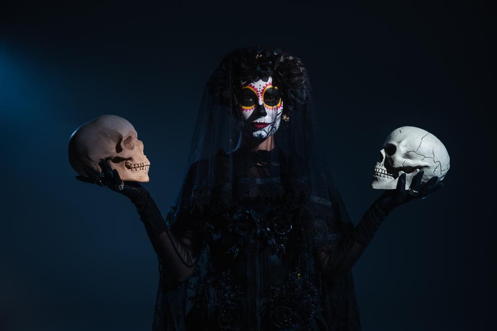 front view of woman in halloween makeup and black costume standing with skulls on dark blue background - Photo, Image