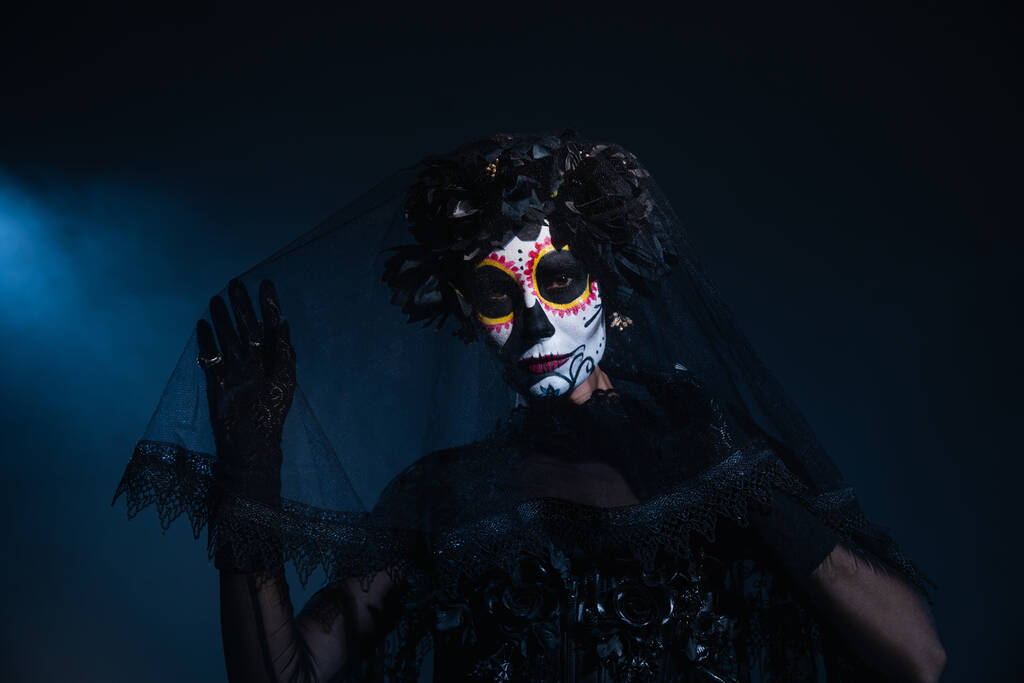 mulher na maquiagem tradicional santa muerte tocando véu preto e olhando para a câmera no fundo escuro com luz azul - Foto, Imagem