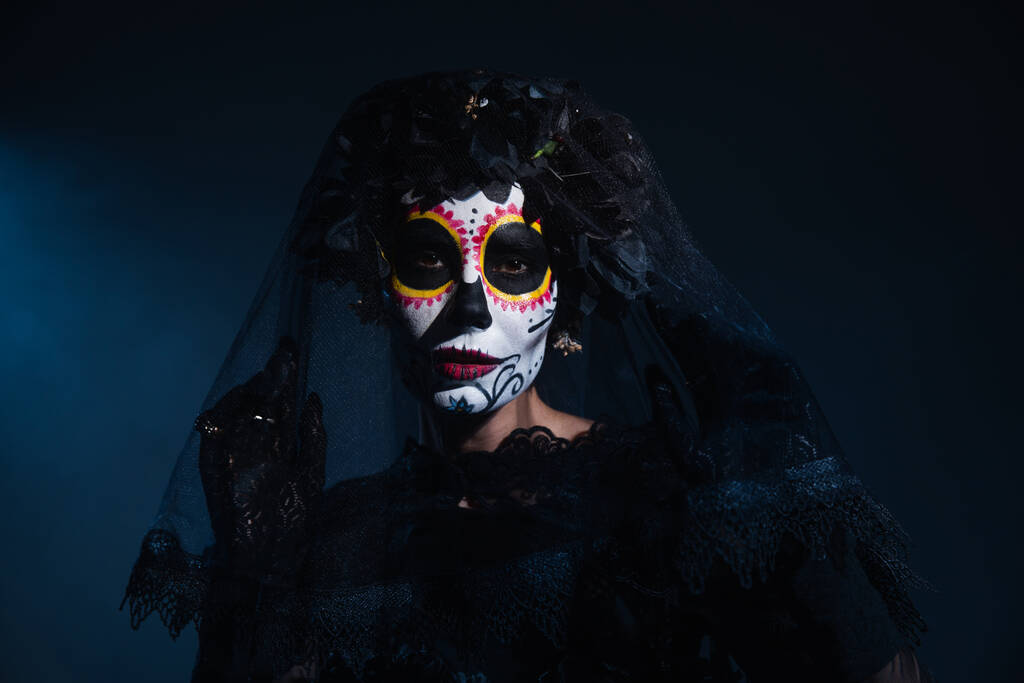 женщина в мексиканском дне мертвого костюма и грима Катрины, смотрящая на камеру под черным кружевным покрывалом на темно-синем фоне - Фото, изображение