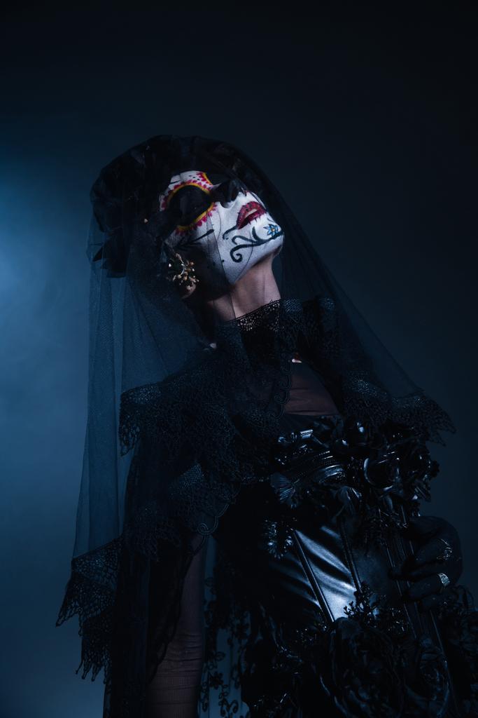 woman in creepy santa muerte makeup and black costume posing with closed eyes on black and blue background - Photo, Image